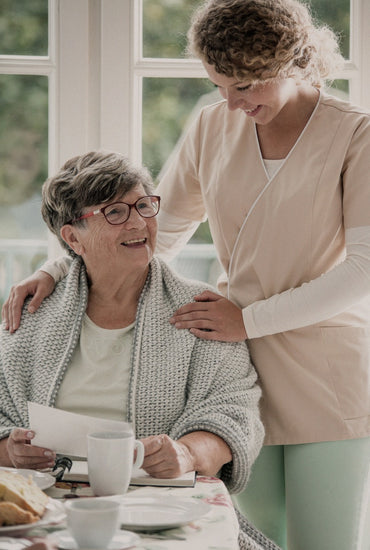 Eine ältere Frau sitzt, ihr Betreuer steht neben ihr, beide sehen glücklich aus.